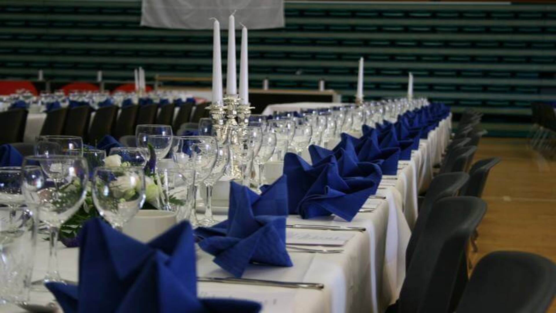 Bordopdækning til fest i Hal 1. Service med blå servietter, hvide stearinlys og blomster buketter til pyntning.